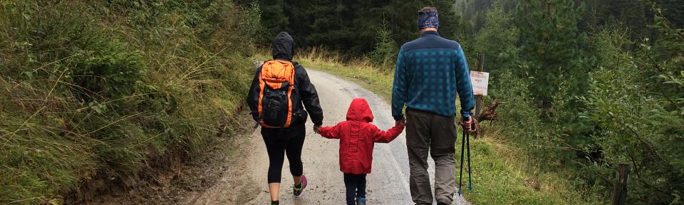 Two parents walking on either side of a child on a trail. The parent on the left is wearing a black jacket with an orange backpack, and holding one hand of the child who is wearing a red rain coat. The parent on the right is holding the child's other hand and is wearing a blue plaid jacket and tan pants. They are hiking up a trail surrounded by green foliage